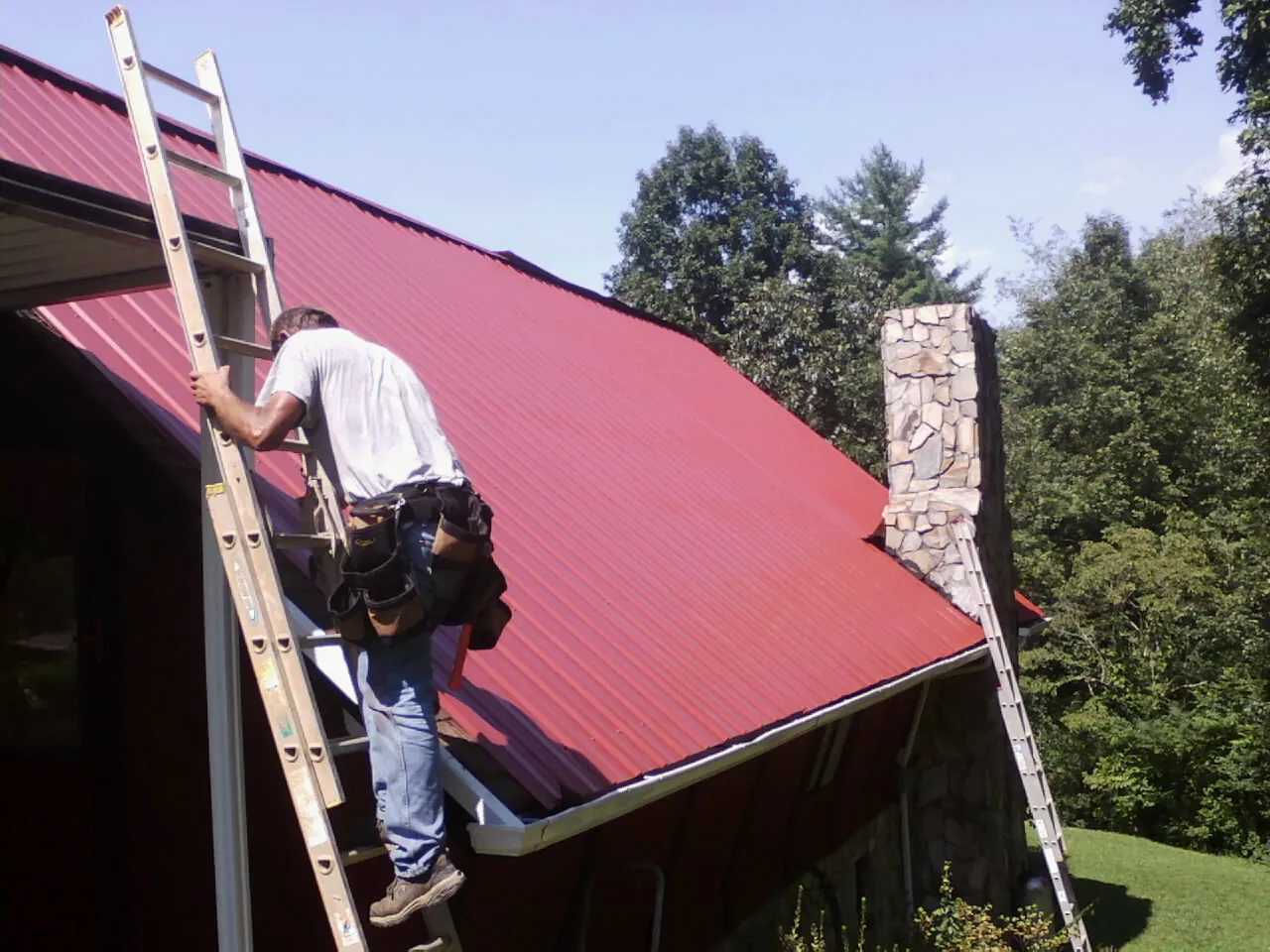 Roofer on a ladder beside a red metal roof and stone chimney — Soffit & Fascia Repair by Roofing Brothers in Langston